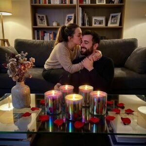 Girl kissing boyfriend on forehead and hugging him for attention, five Lucentius rainbow glass candles on glass table with red rose petals scattered around, romantic atmosphere